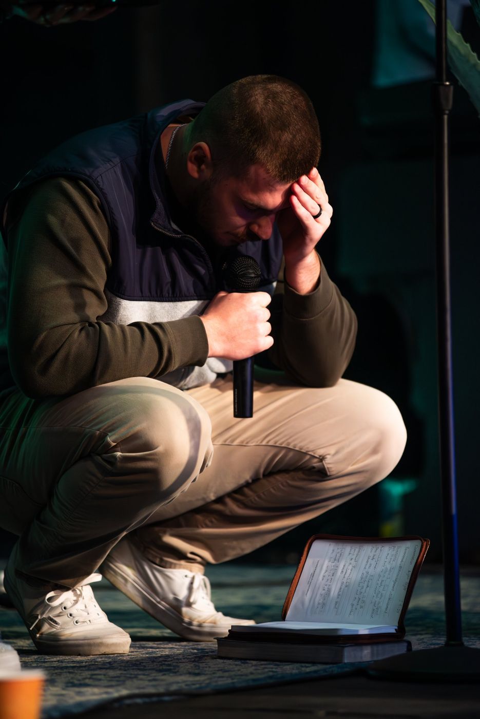 A young man crouched low in prayer, microphone in hand, his Bible and handwritten notes open on the floor