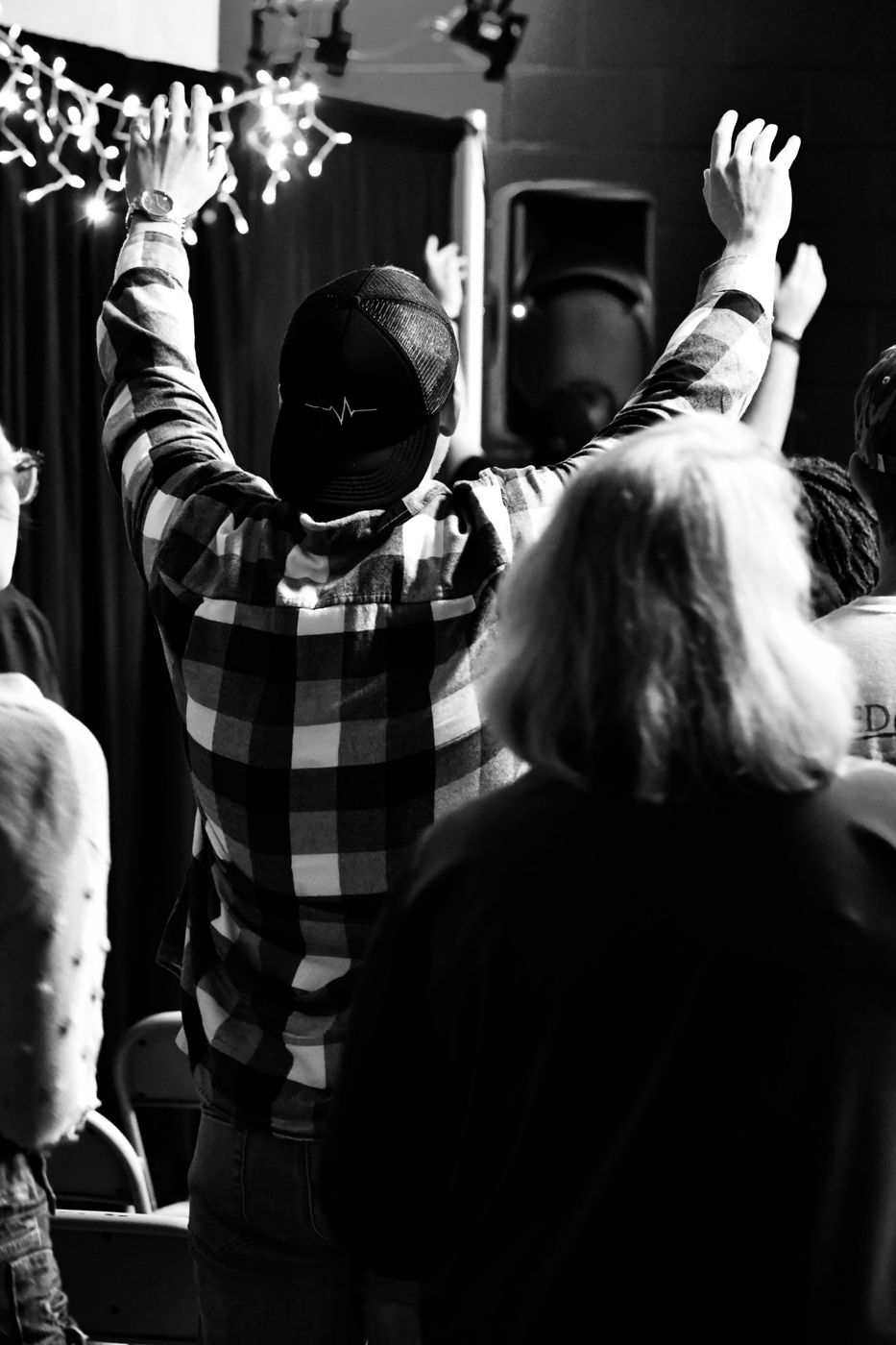 A man in a flannel shirt and cap with both arms raised under string lights