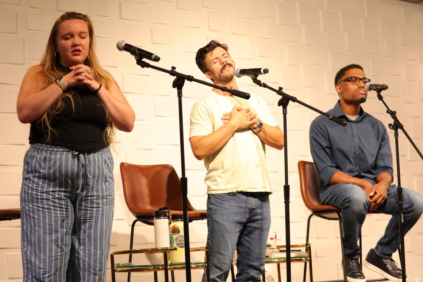 Three people praying at microphones, eyes closed, hands on their hearts